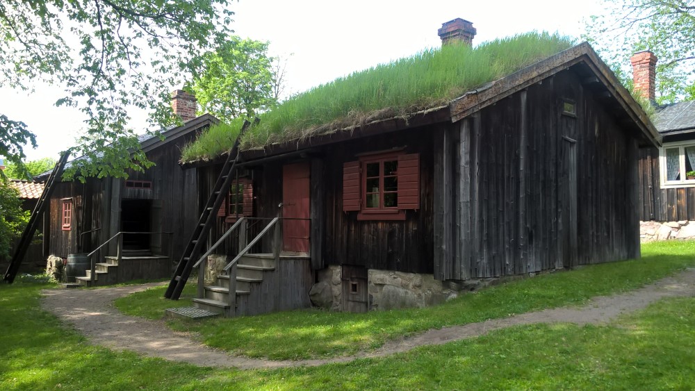 Ruohokattoinen maalaamaton puurakennus, jossa puneiset ikkunapuut ja ovi. Edessä portaat ovelle.