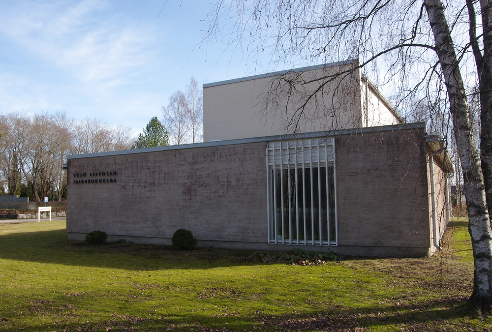 Betoninen harmaa rakennus, jonka oikeassa reunassa on pystyrimainen ikkuna ja vasemmassa reunassa teksti Yrjö Liipolan taidemuseo. Museon edessä on nurmikenttä.