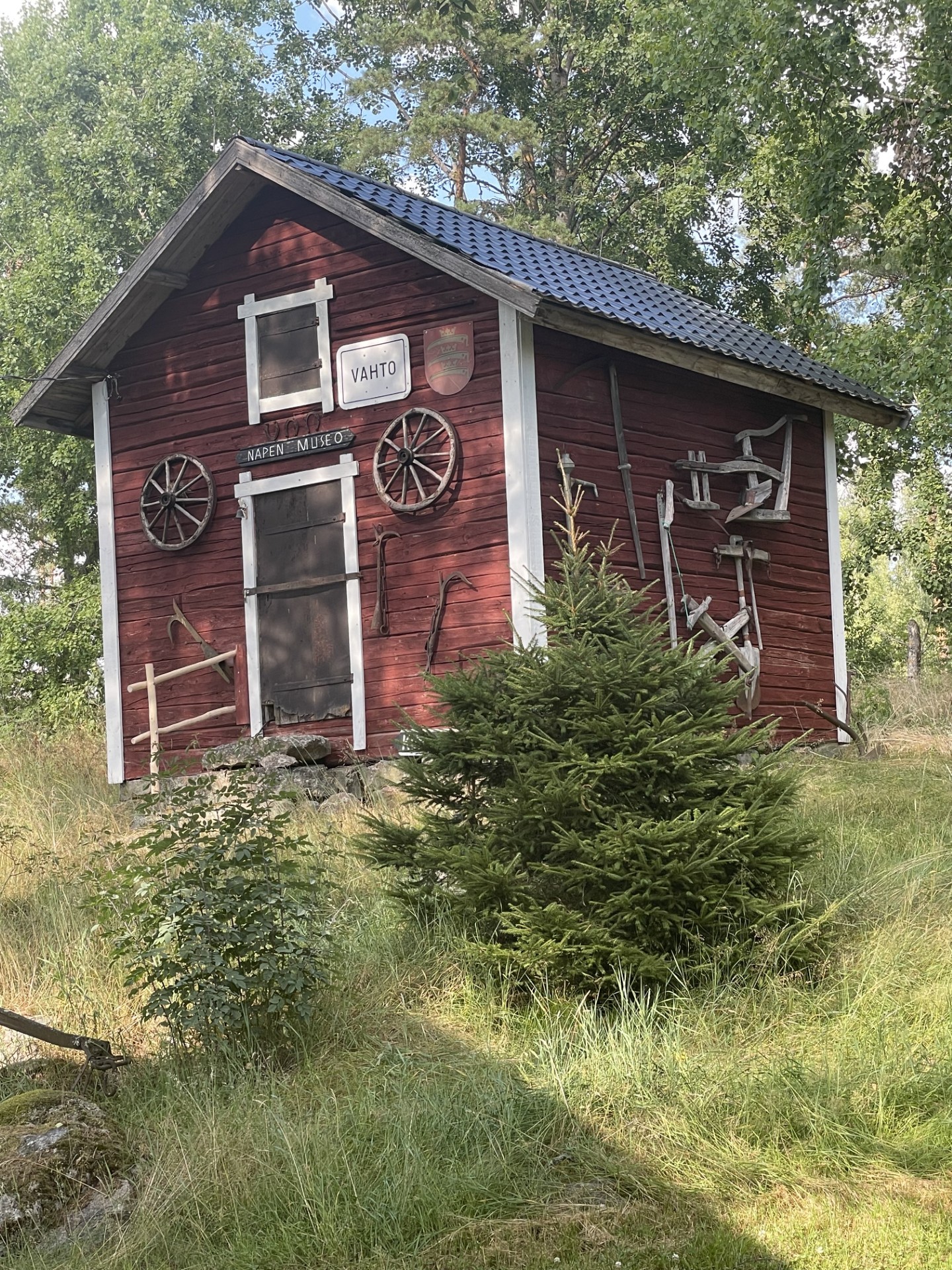 Punainen pieni rakennus, jossa edessä ovi ja sen molemmin puolin kärrynpyörät. Toisella seinällä lisää puisia maatalousvälineitä.
