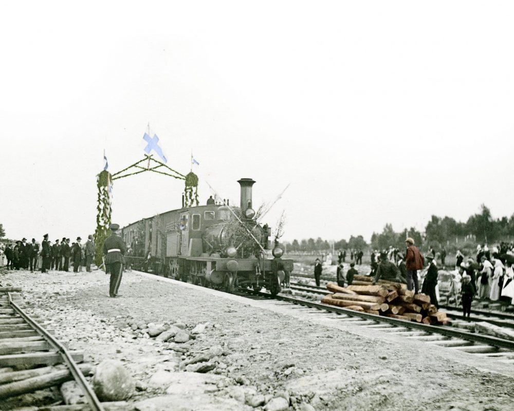Ensimmäinen juna Uudessakaupungissa 1924. Mustavalkoinen kuva, jossa edessä raiteet, raiteiden vieressä ihmisiä, raiteilla kulkee höyryjuna koristeellisen portin läpi.